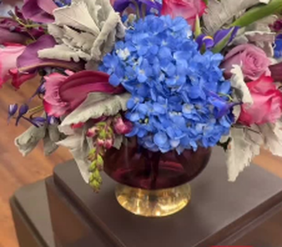 Blue hydrangea arrangement with pink accents in a footed gold and glass vase